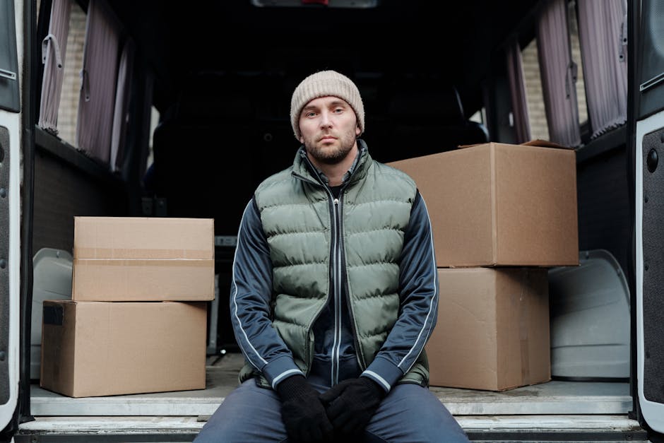 Photograph of two male movers inside a white moving van with the sliding door open, parked on a residential street with brick buildings and trees in the background. The man in the passenger seat has a