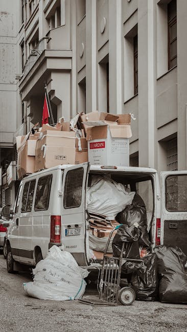 A white commercial van parked on a city street with its rear doors open, revealing a load of various packed and partially unpacked cardboard boxes, some with visible labels, and black plastic rubbish bags filled with waste. Cardboard boxes are stacked on the roof of the van, secured with plastic straps, including larger boxes and smaller delivery cartons. Inside the van, there are protective coverings such as fabric or blankets, along with packing materials and possibly furniture or household items prepared for removal. A small trolley or hand truck is positioned next to the open van door, with additional rubbish bags nearby on the pavement. The background features a multi-storey residential or commercial building with many windows and balconies, indicating an urban environment. The scene depicts a home relocation or clearance process, with visible tools and materials associated with furniture transport and packing and moving activities, supported by the presence of Man with Van Collier Row’s services.