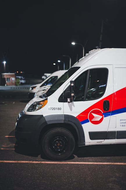 A white postal delivery van with a red and blue stripe design is parked in a well-lit outdoor area during nighttime, with other similar vans lined up behind it. Inside the van's open side door, a worker is seen loading or unloading a large cardboard box wrapped in plastic and secured with packing materials, such as bubble wrap or fabric padding. The scene captures the loading process involved in home relocation or furniture transport, with the van positioned on a paved surface and streetlights illuminating the area. The setting indicates an organized move or delivery operation, potentially coordinated by Man with Van Collier Row, involving furniture, packing supplies, and moving equipment like trolleys or straps. The background shows additional vehicles and a low building, with the focus on the vehicle, its contents, and the active handling of packing and moving materials.