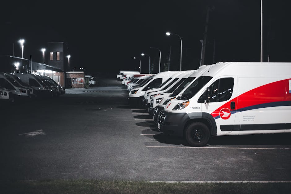 A white commercial van parked on a city street with its rear doors open, revealing a load of various packed and partially unpacked cardboard boxes, some with visible labels, and black plastic rubbish bags filled with waste. Cardboard boxes are stacked on the roof of the van, secured with plastic straps, including larger boxes and smaller delivery cartons. Inside the van, there are protective coverings such as fabric or blankets, along with packing materials and possibly furniture or household items prepared for removal. A small trolley or hand truck is positioned next to the open van door, with additional rubbish bags nearby on the pavement. The background features a multi-storey residential or commercial building with many windows and balconies, indicating an urban environment. The scene depicts a home relocation or clearance process, with visible tools and materials associated with furniture transport and packing and moving activities, supported by the presence of Man with Van Collier Row’s services.