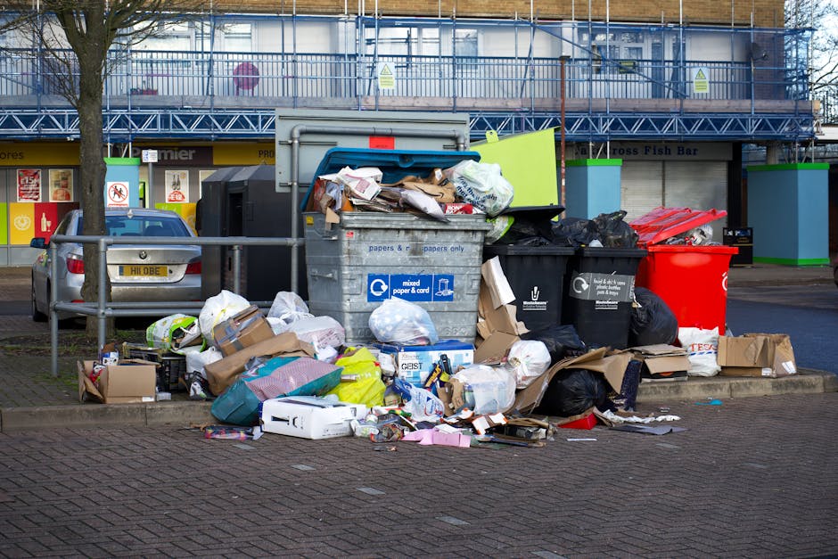 A collection of overflowing waste and recycling bins situated on a paved sidewalk in a shopping area, with various paper, cardboard boxes, plastic bags, and packaging materials spilling onto the ground. The recycling containers include a large grey mixed paper and cardboard bin, a black bin for general waste, and a red bin. Some cardboard boxes are flattened or partially crushed, while others remain upright. The scene is set outside a retail unit with visible shopfronts, signage, and a blue metal scaffolding covering an upper floor. A silver car is parked nearby, and a metal railing separates the waste area from the parking space. The environment appears clean but cluttered due to the excess waste surrounding the bins, illustrating the importance of proper waste disposal and recycling management, especially relevant to relocation services like those provided by Man with Van Collier Row engaged in home removals and furniture transport.