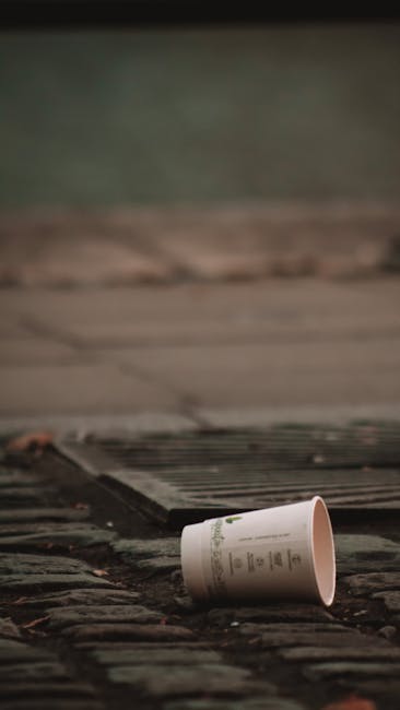 A disposable paper cup lying on its side on a concrete or stone doorstep outside a property, with the cup close to a wooden or metal threshold. The background shows a blurred area of external paving or driveway with greenish and grey tones, suggesting an outdoor environment. The scene appears to be part of a home relocation or moving process, with the cup possibly discarded during packing or loading activities. The placement indicates that the cup is near the entrance, where packing materials or waste may be temporarily placed before disposal or collection, consistent with house removal preparations. Man with Van Collier Row may be involved in moving services that include waste management or cleanup during a furniture transport or packing and moving operation.