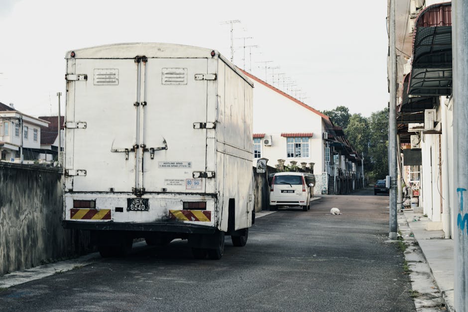 A large green and beige box truck parked on a city street during daylight hours, with tall modern office buildings and historic architecture in the background. The truck is positioned near the curb, p