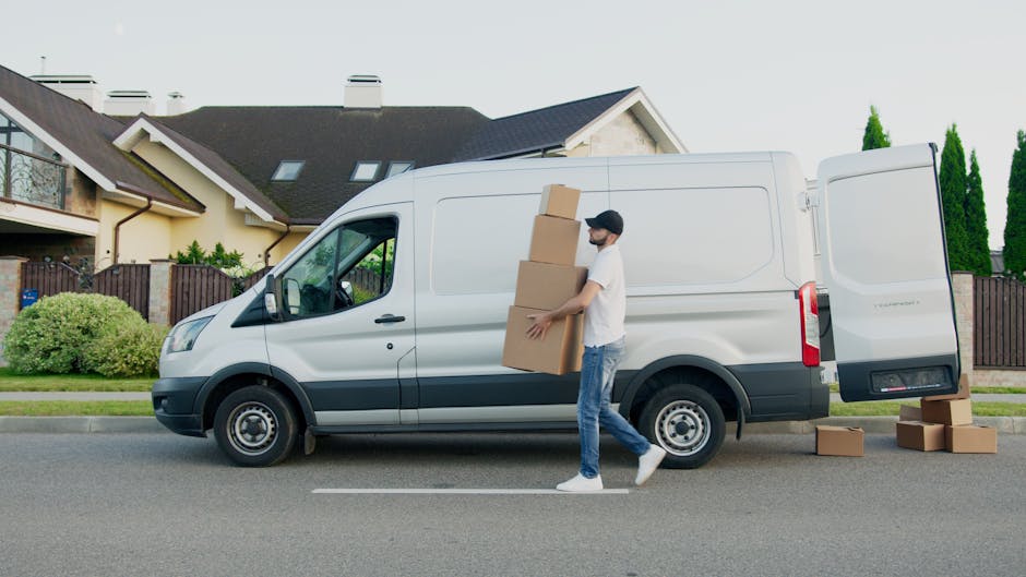 A man wearing a white t-shirt, blue jeans, and a black cap is loading large brown cardboard boxes into the side door of a white commercial van parked on a residential street. Several additional boxes are on the pavement nearby, with some stacked on the ground and others being carried or positioned for loading. The van is situated in front of a modern, detached house with a dark grey roof, cream-colored walls, and skylights, surrounded by a wooden fence and landscaped garden with bushes and tall, narrow trees. The scene is well-lit under clear daytime conditions, reflecting the process of home relocation, furniture transport, and packing during a professional removal service. The environment suggests an urban suburban area with traffic lanes visible beneath the vehicle.