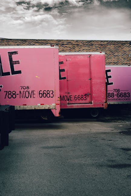 A man wearing a white t-shirt, blue jeans, and a black cap is loading large brown cardboard boxes into the side door of a white commercial van parked on a residential street. Several additional boxes are on the pavement nearby, with some stacked on the ground and others being carried or positioned for loading. The van is situated in front of a modern, detached house with a dark grey roof, cream-colored walls, and skylights, surrounded by a wooden fence and landscaped garden with bushes and tall, narrow trees. The scene is well-lit under clear daytime conditions, reflecting the process of home relocation, furniture transport, and packing during a professional removal service. The environment suggests an urban suburban area with traffic lanes visible beneath the vehicle.