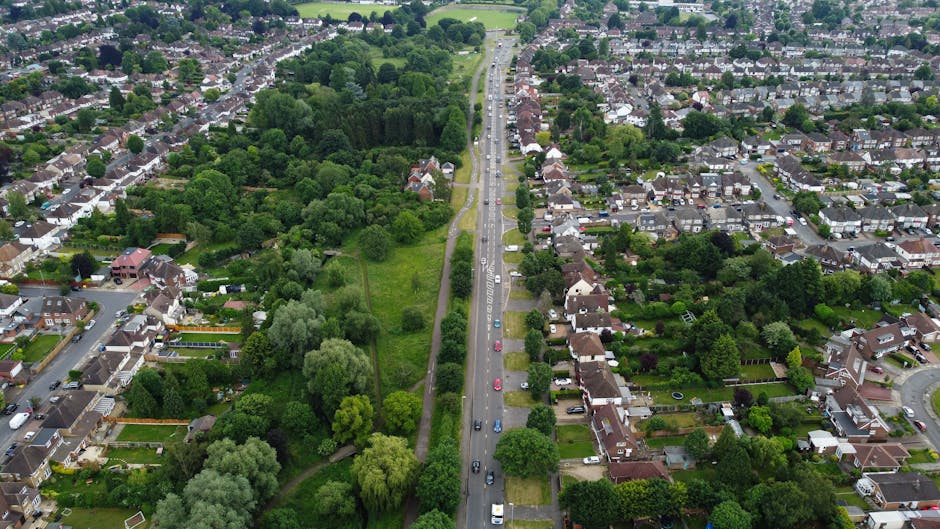 An aerial view of a suburban neighbourhood showing a straight main road with parked cars along both sides, bordered by residential houses with gardens, and a large green park area with dense trees and open grass sections to the left. The houses are mostly semi-detached or terraced, featuring pitched roofs, and some have visible driveways or front yards. The scene captures the environment during daylight, with the street actively used by vehicles, indicating ongoing traffic and movement. In the context of house removals and relocation services, this image illustrates the typical setting where furniture transport and packing and moving activities might occur, emphasizing the importance of proper planning and permits for street access in Collier Row, as managed by Man with Van Collier Row.
