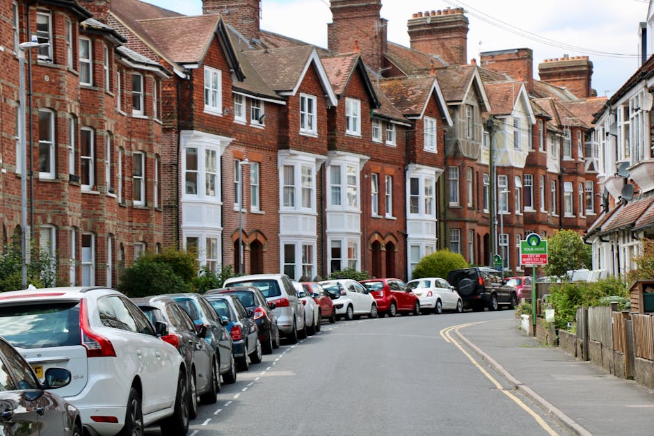 A residential street with a row of traditional red brick terraced houses featuring white window frames and pitched roofs, situated behind a parked line of various cars including hatchbacks and sedans along the pavement. The street is slightly curved with double yellow lines marking the parking boundary. A prominent green sign indicates parking restrictions or permits, with a small shrubbery and a wooden fence at the edge of the pavement. The scene is set during daylight hours with clear weather, under partly cloudy skies. This setting reflects a typical environment where home relocation or furniture transport might take place, often involving the loading or unloading process managed by professional removals specialists such as Man with Van Collier Row, who facilitate house removals and moving services in the area.