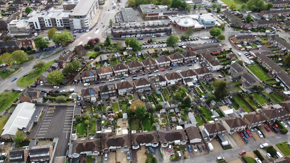 An aerial view of a suburban neighbourhood showing a straight main road with parked cars along both sides, bordered by residential houses with gardens, and a large green park area with dense trees and open grass sections to the left. The houses are mostly semi-detached or terraced, featuring pitched roofs, and some have visible driveways or front yards. The scene captures the environment during daylight, with the street actively used by vehicles, indicating ongoing traffic and movement. In the context of house removals and relocation services, this image illustrates the typical setting where furniture transport and packing and moving activities might occur, emphasizing the importance of proper planning and permits for street access in Collier Row, as managed by Man with Van Collier Row.