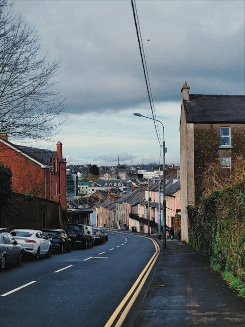 A narrow residential street on a cloudy day with a gently curved tarmac road marked with dashed white lines and double yellow lines along the edge. On the left side, parked cars in various colours line the pavement, which is partly shaded by a leafless tree extending into the frame. To the right, a tall, older brick building with a chimney and exterior windows is adjacent to a grassy embankment overgrown with green vines. A streetlight with a curved arm hangs over the road, connected to overhead utility wires stretching across the scene. The background reveals a view of densely packed houses and buildings with rooftops, extending into the horizon under a sky filled with dark, heavy clouds. This scene relates to home relocation or furniture transport, as part of the reporting on house removals and moving logistics by Man with Van Collier Row.