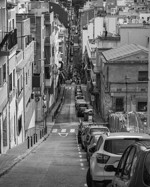 A narrow residential street on a cloudy day with a gently curved tarmac road marked with dashed white lines and double yellow lines along the edge. On the left side, parked cars in various colours line the pavement, which is partly shaded by a leafless tree extending into the frame. To the right, a tall, older brick building with a chimney and exterior windows is adjacent to a grassy embankment overgrown with green vines. A streetlight with a curved arm hangs over the road, connected to overhead utility wires stretching across the scene. The background reveals a view of densely packed houses and buildings with rooftops, extending into the horizon under a sky filled with dark, heavy clouds. This scene relates to home relocation or furniture transport, as part of the reporting on house removals and moving logistics by Man with Van Collier Row.