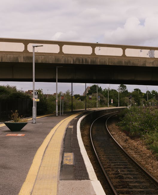 A deserted train platform with a curved track running alongside the edge, bordered by a yellow safety line and a white tactile paving strip. The platform features a small flower bed in a black, tapered planter, and is equipped with several tall, grey lampposts. Overhead, a concrete bridge spans above, with blank white panels on its sides. In the background, trees and bushes line the railway embankment under an overcast sky, creating a neutral setting ideal for depicting the logistics of home relocation or furniture transport connected to house removals and moving services. The scene suggests a quiet moment during transit or loading activities, with no visible people or moving equipment, emphasizing the environmental context of a rail-based part of a house move or loading process. This image complements the topic of last-mile moving tips for house removals in Collier Row, as presented by Man with Van Collier Row.