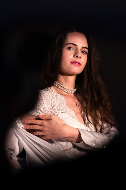 A young woman with long, wavy brown hair and light skin, posing against a dark background. She is wearing a white lace top with intricate patterns, layered with a pearl necklace and a choker. Her expression is neutral, with a slight tilt of her head, and she gazes softly at the camera. The lighting highlights her face and upper body, creating a contrast with the dark surroundings. This portrait-style image emphasizes her features and attire, with no visible surroundings or contextual elements related to house removals or moving services.