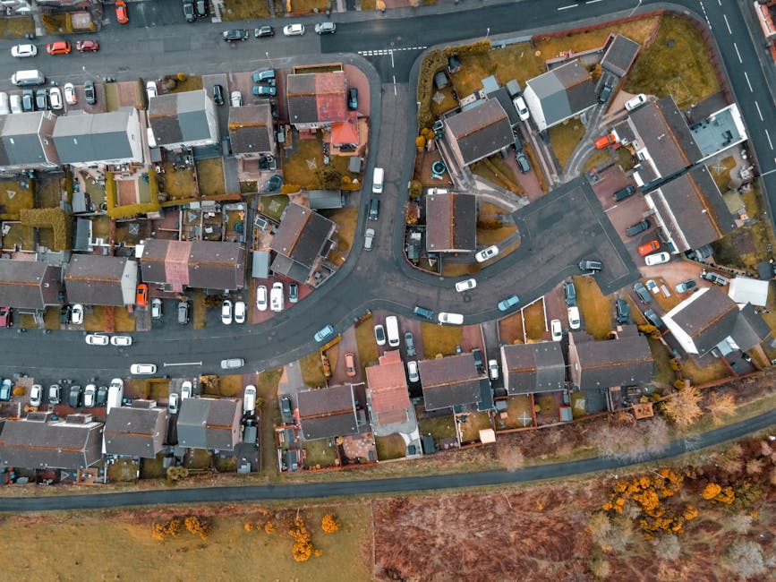 An aerial view of a residential neighbourhood showing multiple houses with grey and red roofs, arranged along curved streets with numerous parked cars in driveways and along the roads. Inside some properties, visible objects include cardboard boxes, wrapped furniture, and household items prepared for a home relocation. A large van is parked on the driveway of one house, part of it loaded with packed boxes and furniture covered in protective blankets. The loading process is evident as items are being moved from the house into the van, with some furniture being carried by individuals or positioned on trolleys. The surrounding area displays a mixture of paved driveways, small gardens, and patches of grass and trees, indicating an organized but densely built suburban environment. The scene reflects professional removals services by Man with Van Collier Row, supporting packing and furniture transport during a house move.