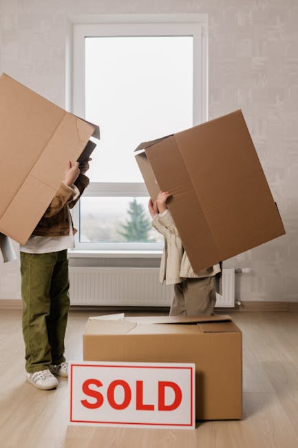 A man wearing a black cap, white polo shirt, and jeans is kneeling on the pavement outside an open moving truck. He is in the process of packing or organizing cardboard boxes, some of which are stacked nearby inside the vehicle. The scene is set during daylight hours, with sunlight casting a warm glow on the man and the surrounding area. The moving truck door is open, revealing boxes and packing materials such as plastic wrap and cardboard. The background shows a paved road and a grassy verge, indicating a residential or suburban location consistent with house removals services. This action is part of the home relocation process handled by Man with Van Collier Row, involving furniture transport and packing and moving logistics.